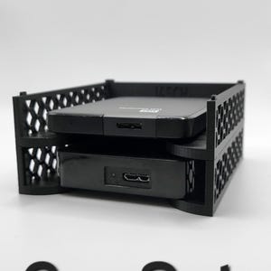 Pode incluir: Dois discos rígidos externos pretos em uma caixa de armazenamento de plástico preto. A caixa tem um padrão de grade e o texto "2er Set" é impresso abaixo da caixa.