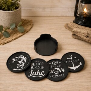 May include: A set of four circular black coasters with white graphic designs featuring fishing themes, including a fish illustration, text reading Life is better at the lake, Eat Sleep Fish Repeat, and Petri heil with an anchor, plus a black holder.