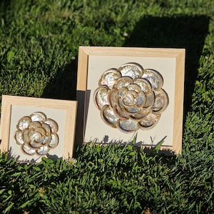May include: Two framed wall art pieces featuring a flower design made from iridescent seashells. The larger frame is light brown wood with a white mat and the smaller frame is a darker brown wood with a white mat.