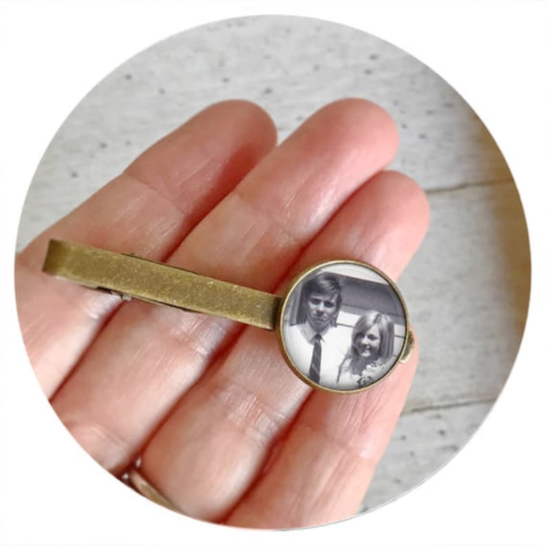May include: A bronze tie clip with a round glass insert featuring a black and white photograph of a couple. The man is wearing a suit and tie, and the woman is wearing a dress.