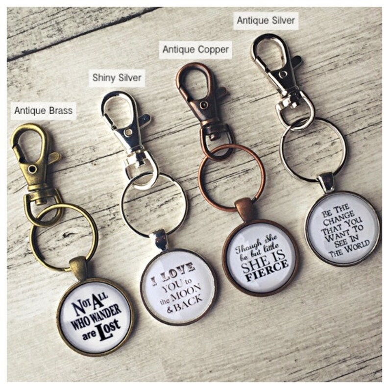 May include: Five keychains with different finishes: antique brass, shiny silver, antique copper, and antique silver. Each keychain has a round charm with a different inspirational quote. The charms read: "Not All Who Wander Are Lost", "I Love You To The Moon & Back", "Though She Be But Little She Is Fierce", and "Be The Change That You Want To See In The World".