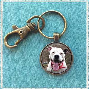 May include: A bronze key chain with a round charm featuring a white and black dog with a red collar. The dog is smiling with its tongue sticking out.