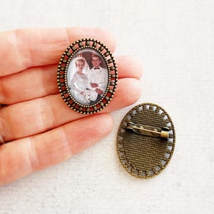 May include: Oval-shaped vintage brooch with a sepia-toned wedding photo. The brooch has a bronze-colored frame with a beaded edge. The back of the brooch has a pin closure. The image shows a bride and groom.