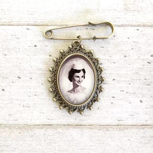 May include: A bronze-colored brooch with a safety pin closure. The brooch features an oval-shaped frame with an ornate design. Inside the frame is a black and white photo of a woman in a wedding dress.
