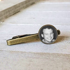 May include: A gold tie bar with a round, black and white photo of a man smiling. The photo is set in a clear, round, glass-like casing.