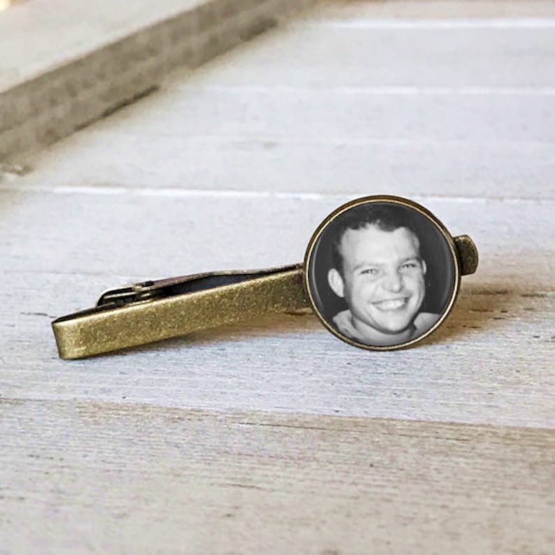 May include: A gold tie bar with a round, black and white photo of a man smiling. The photo is set in a clear, round, glass-like casing.
