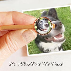 May include: A close-up of a custom tie tack featuring a photo of a black and white dog. The tie tack is made of metal and has a round, clear glass dome. The dog is looking directly at the camera with its mouth open in a smile.