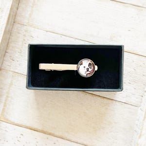 May include: A silver tie clip with a round, clear glass cabochon featuring a black and white dog's face. The tie clip is in a black velvet box.