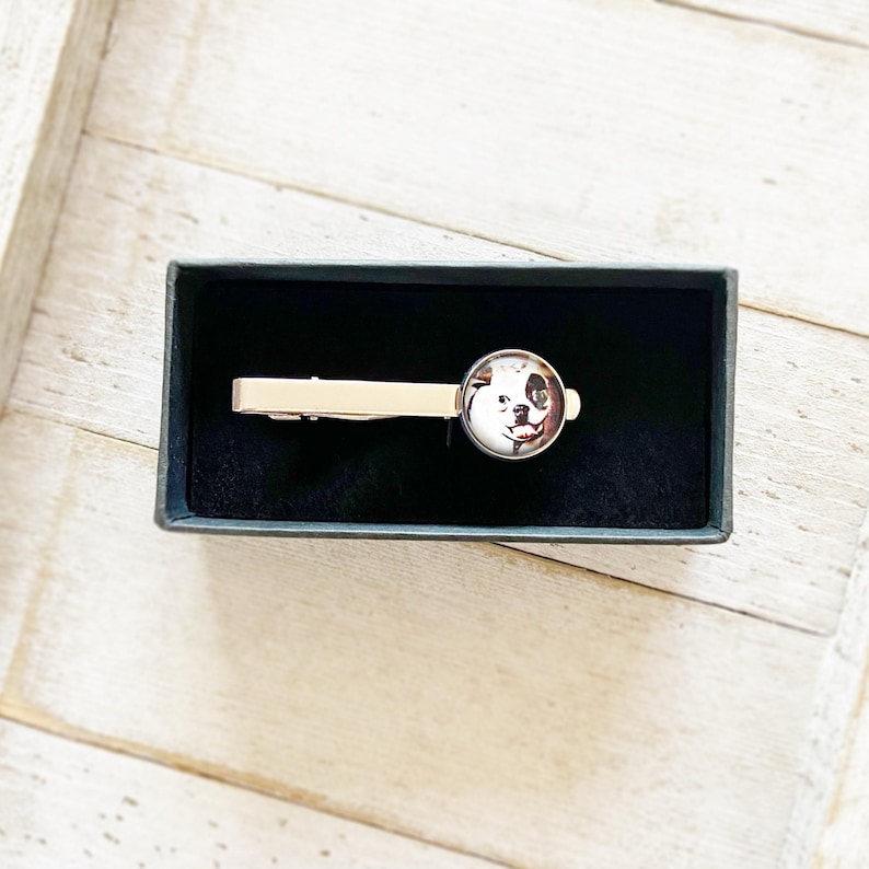 May include: A silver tie clip with a round, clear glass cabochon featuring a black and white dog's face. The tie clip is in a black velvet box.