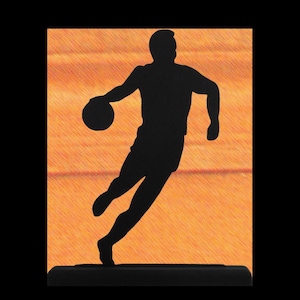 May include: A black silhouette of a basketball player dribbling a ball, set against an orange textured background. The figure is mounted on a black base, creating a dynamic sports-themed design.