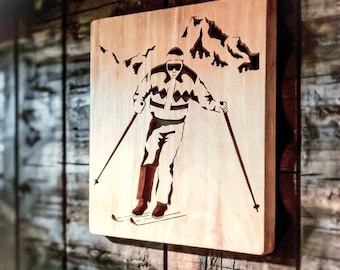 Op de afbeelding: Een vierkante houten wandplaat met een donkerbruin lasergesneden of gegraveerd ontwerp van een skiër in beweging. De skiër draagt een skibril en een trui met patroon en houdt twee skistokken vast voor een afgebeeld berglandschap.