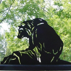 Peut inclure: Silhouette de métal noir d'un ours assis sur un rocher. L'ours est tourné vers la gauche et présente un design détaillé.