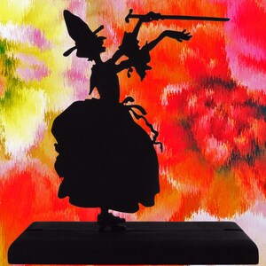 Könnte beinhalten: Silhouette einer Hexe, die ein Schwert hält und auf einem schwarzen Holzsockel vor einem bunten Hintergrund steht.