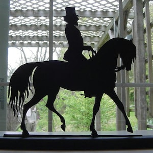 Pode incluir: Silhueta preta de uma pessoa a cavalo. A silhueta é cortada em metal e montada em uma base preta.