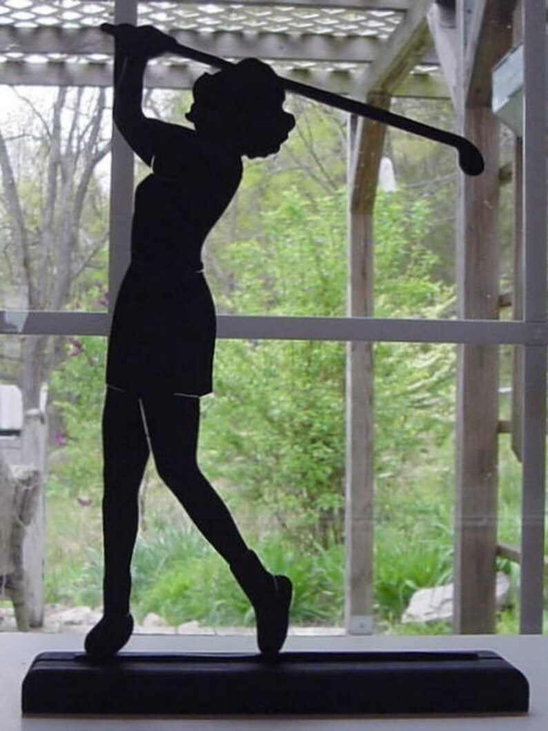 Puede incluir: Silueta negra de una golfista en pleno swing, sosteniendo un palo de golf. La figura est&aacute; sobre una base rectangular. El fondo muestra una ventana con vistas a &aacute;rboles y vegetaci&oacute;n. Un objeto decorativo.