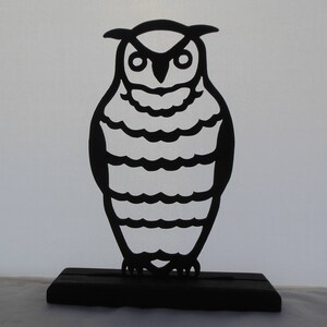 Könnte beinhalten: Schwarze Silhouette einer Eule mit einem Holzsockel. Die Eule hat einen runden Körper mit einem gestreiften Muster und großen Augen.