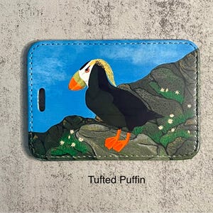 K&ouml;nnte beinhalten: Ein Gep&auml;ckanh&auml;nger mit einer farbenfrohen Illustration eines Papageientauchers auf einer felsigen Landschaft. Der Vogel hat schwarzes Gefieder, orangefarbene F&uuml;&szlig;e und einen gelb-wei&szlig;en Kopf. Der Hintergrund ist blau mit wei&szlig;en Blumen.