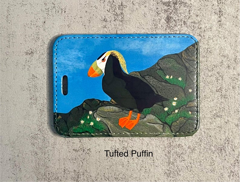 K&ouml;nnte beinhalten: Ein Gep&auml;ckanh&auml;nger mit einer farbenfrohen Illustration eines Papageientauchers auf einer felsigen Landschaft. Der Vogel hat schwarzes Gefieder, orangefarbene F&uuml;&szlig;e und einen gelb-wei&szlig;en Kopf. Der Hintergrund ist blau mit wei&szlig;en Blumen.