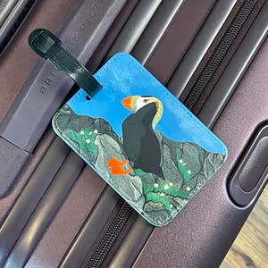 K&ouml;nnte beinhalten: Blaues Gep&auml;ckanh&auml;nger mit einer Illustration eines Papageientauchers. Das Etikett hat einen schwarzen Riemen und befindet sich auf einem strukturierten Hintergrund. Der Papageientaucher ist mit orangefarbenen F&uuml;&szlig;en und Schnabel sowie schwarzen und wei&szlig;en Federn dargestellt.