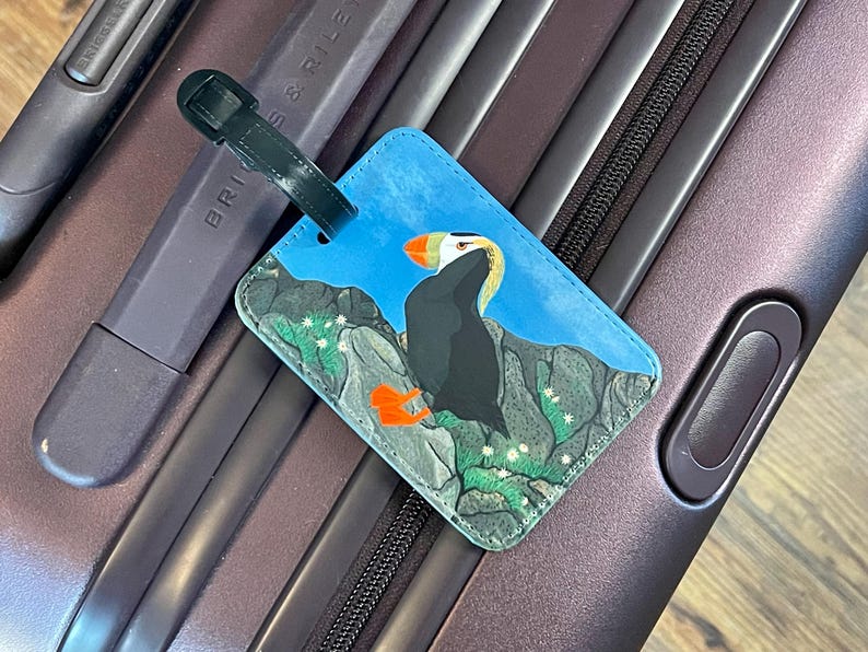 K&ouml;nnte beinhalten: Blaues Gep&auml;ckanh&auml;nger mit einer Illustration eines Papageientauchers. Das Etikett hat einen schwarzen Riemen und befindet sich auf einem strukturierten Hintergrund. Der Papageientaucher ist mit orangefarbenen F&uuml;&szlig;en und Schnabel sowie schwarzen und wei&szlig;en Federn dargestellt.
