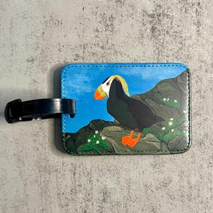 K&ouml;nnte beinhalten: Rechteckiger Gep&auml;ckanh&auml;nger mit einer Illustration eines Papageientauchers. Der Anh&auml;nger zeigt einen Papageientaucher auf einem Felsen mit blauem Himmel im Hintergrund. Er hat einen schwarzen Riemen zur Befestigung. Der Papageientaucher hat orange F&uuml;&szlig;e und einen Schnabel.