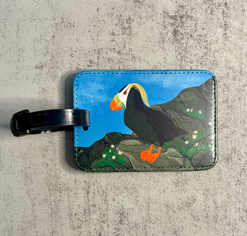 K&ouml;nnte beinhalten: Rechteckiger Gep&auml;ckanh&auml;nger mit einer Illustration eines Papageientauchers. Der Anh&auml;nger zeigt einen Papageientaucher auf einem Felsen mit blauem Himmel im Hintergrund. Er hat einen schwarzen Riemen zur Befestigung. Der Papageientaucher hat orange F&uuml;&szlig;e und einen Schnabel.