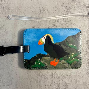 K&ouml;nnte beinhalten: Ein Gep&auml;ckanh&auml;nger mit einer Illustration eines Papageientauchers. Der Anh&auml;nger hat einen blauen Hintergrund mit einem Papageientaucher auf einem Felsen. Der Papageientaucher hat einen gelb-orangen Schnabel und orangefarbene F&uuml;&szlig;e. Ein schwarzer Riemen ist ebenfalls vorhanden.