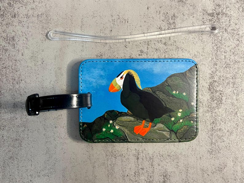 K&ouml;nnte beinhalten: Ein Gep&auml;ckanh&auml;nger mit einer Illustration eines Papageientauchers. Der Anh&auml;nger hat einen blauen Hintergrund mit einem Papageientaucher auf einem Felsen. Der Papageientaucher hat einen gelb-orangen Schnabel und orangefarbene F&uuml;&szlig;e. Ein schwarzer Riemen ist ebenfalls vorhanden.