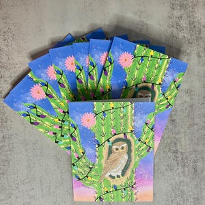 Puede incluir: Un conjunto de tarjetas de felicitación con un diseño de cactus caprichoso. Cada tarjeta muestra un cactus verde con flores rosas, luces de cadena y un búho. El fondo es un degradado de azul y morado.