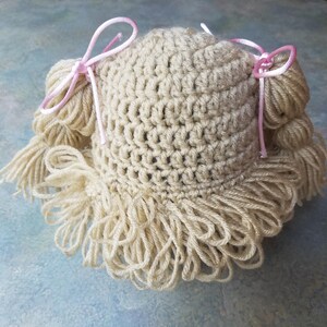 May include: A handmade, off-white crochet doll wig with long, fringed hair and two pink ribbon bows. The wig is designed to fit a doll's head and is made of soft yarn. The crochet stitches are visible, adding to the handcrafted appeal.