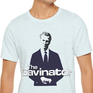 May include: Light blue t-shirt featuring a navy blue silhouette of a man in a suit with the text "The Gavinator" in white. The shirt is worn by a person.