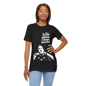 May include: A black short-sleeve t-shirt featuring a white graphic print of a person with their tongue out, wearing a star-patterned top. Above the graphic, the text reads: The fear Addicted, A danger illustrated. The shirt is paired with blue jeans.