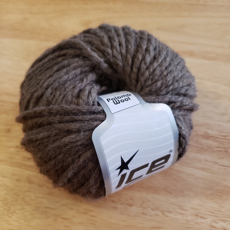 May include: A ball of brown Paloma Wool yarn with a white label featuring the brand's logo. The yarn is thick and textured, suitable for knitting or crocheting projects. The ball of yarn is resting on a wooden surface.