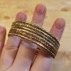 May include: A set of gold-toned bangles with a textured, hammered finish. The bangles are stacked on top of each other, creating a layered look.