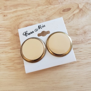 May include: A pair of round, cream-colored stud earrings with a gold-tone metal border. The earrings are displayed on a white card with the text "Casa Mia" in cursive script.