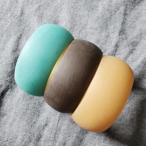 May include: Three wooden bangles in shades of teal, brown, and tan. The bangles are smooth and curved, with a slightly rounded rectangular shape. The colors are solid and the bangles are stacked together.