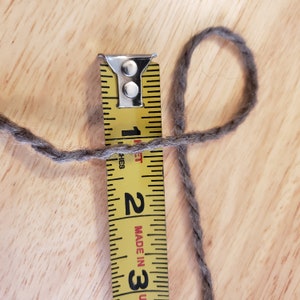 May include: A close-up shot shows a gray yarn loop next to a yellow measuring tape. The tape reads approximately 1 inch. The tape is marked with black numbers and lines, and the yarn is a neutral gray color. The background is a light wood surface.
