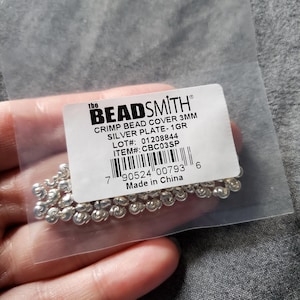 May include: A clear plastic bag containing silver crimp bead covers. The label reads "the BEADSMITH" and includes details like "CRIMP BEAD COVER 3MM SILVER PLATE-1GR" and a barcode. The beads are small and metallic.