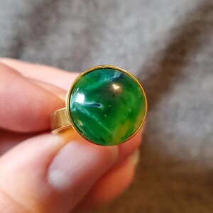 May include: A gold-toned ring featuring a large, round, green cabochon with swirling white and blue patterns. The ring has a simple, classic design, with a smooth, polished finish. The gemstone is set in a gold bezel.