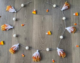 May include: A Halloween-themed garland featuring white, orange, and yellow tassels, white felt balls, and candy corn pieces. The garland is strung on a black and white twine and is displayed on a wooden surface.