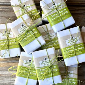May include: A pile of wrapped bars of soap with a green and white label that reads "Small Batch Lanolin Soap" and "Luxurious Lather Naturally Moisturizing" with a sheep illustration. The label also includes the text "Stoney Mountain Farm".