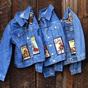 May include: Three blue denim jackets with various cowboy-themed patches. The patches feature cowboys, horses, cacti, and the word "Cowboy".
