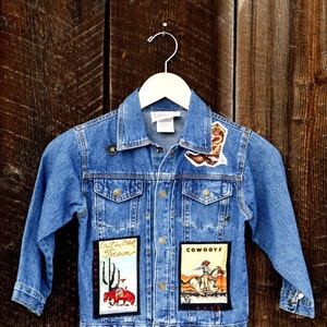 May include: A blue denim jacket with patches. The patches feature images of cowboys, cacti, and the text "Out in Old Tucson" and "COWBOYS".