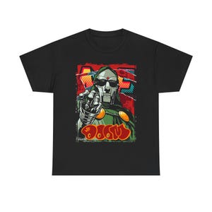 以下が含まれることがあります： 黒いTシャツ。マスク姿の人物が指を指しているグラフィック。緑色のフード、銀色のマスク、落書き風の文字で「MF DOOM」と書かれています。背景は赤、オレンジ、青が混ざっています。