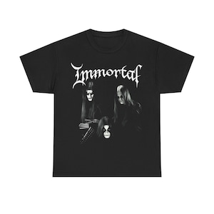 Op de afbeelding: Zwart T-shirt met de band Immortal. De witte afbeelding toont drie figuren met donkere make-up en lang haar. De bandnaam "Immortal" is geschreven in een gestileerd lettertype boven de figuren.