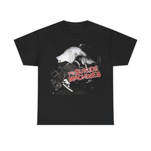 Może przedstawiać: Czarny t-shirt z czarno-białym zdjęciem tłumu. Tekst "THE SUICIDE MACHINES" jest wydrukowany w kolorze czerwonym i białym na przodzie. Koszulka wykonana jest z miękkiego, wygodnego materiału.