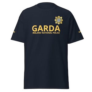 Puede incluir: Camiseta azul marino con la palabra "GARDA" en letras doradas y "IRELAND NATIONAL POLICE" debajo. Un emblema dorado está en la parte superior derecha. La palabra "GARDA" también está impresa en la manga izquierda.
