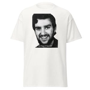 Könnte beinhalten: Ein weißes kurzärmeliges T-Shirt mit einem schwarz-weißen Fotodruck einer lächelnden Person mit Bart. Unter dem Porträt befindet sich der Text Vol. Patsy O' Hara RIP in einer einfachen schwarzen Schrift, mittig auf der Brust platziert.