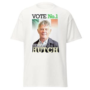 Op de afbeelding: Wit T-shirt met een portret van een man en de tekst "VOTE No.1" erboven en "GERRY HUTCH" eronder. De achtergrond toont de kleuren van de Ierse vlag. Dit is een casual kledingstuk.