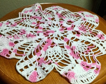 Op de afbeelding: Een handgemaakte, wit en roze gehaakte doily. De doily heeft een bloemenpatroon met openwerk en roze accenten. Het ligt op een houten oppervlak.